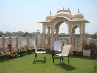 A rooftop terrace with artificial grass features two white chairs and a table in front of a decorative arch structure with domed roofs.