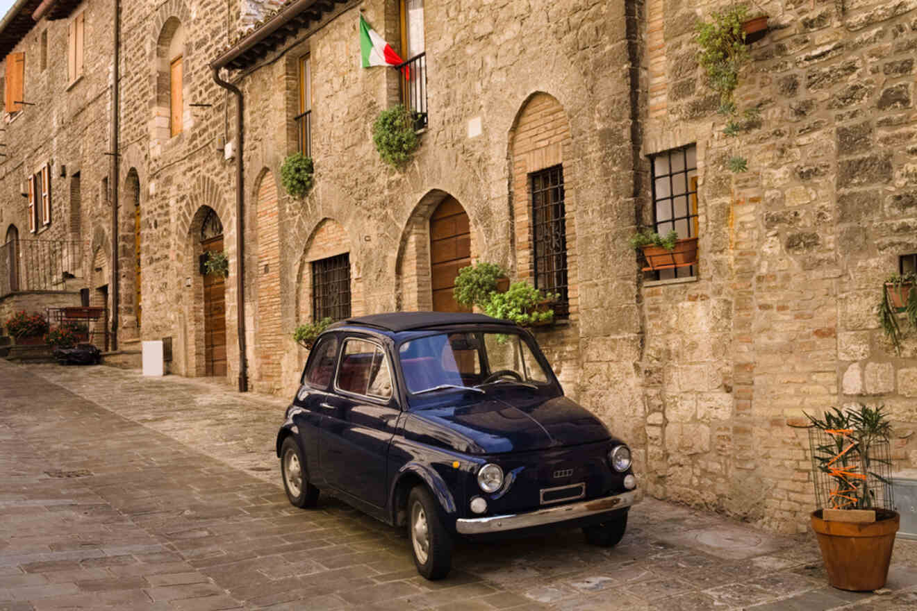 A vintage blue car is parked on a cobblestone street in front of old brick buildings adorned with potted plants and an Italian flag.