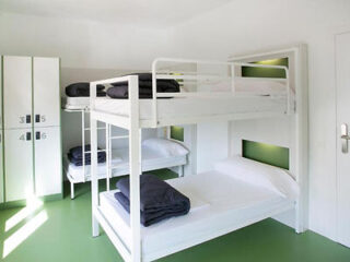 A clean, white room with two sets of metal bunk beds, each with neatly folded black bedding on the top bunks. There are lockers numbered 3 to 6 on the left and a closed white door on the right.