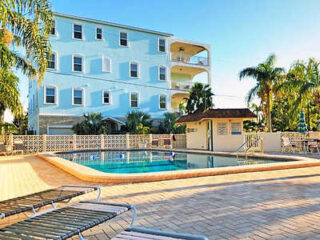 Outdoor pool area of a resort, surrounded by lounge chairs and palm trees, with a light blue hotel building