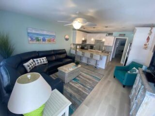 A modern open-plan living room and kitchen featuring a black sectional sofa, teal armchair, bar stools at the kitchen counter, and coastal-themed decor.