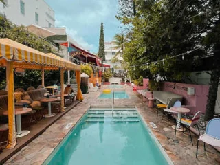 A narrow outdoor pool is flanked by tables, chairs, and striped canopies. The area is adorned with green foliage and string lights, creating a cozy and inviting atmosphere.