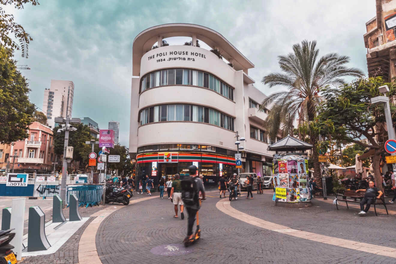 A bustling street scene with a curved white building featuring "The Poli House Hotel" on its facade, palm trees, and people walking or riding scooters around the intersection.