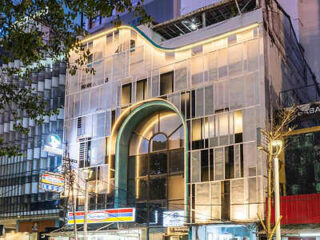 A multi-story building with a uniquely designed facade, featuring an illuminated archway entrance and several retail shops on the ground floor, is shown at dusk.