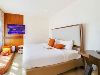 A modern bedroom with a large bed, white linens, and orange pillows. There is a television on the wall, a small seating area with cushions, and a desk in the corner. The room has natural lighting.
