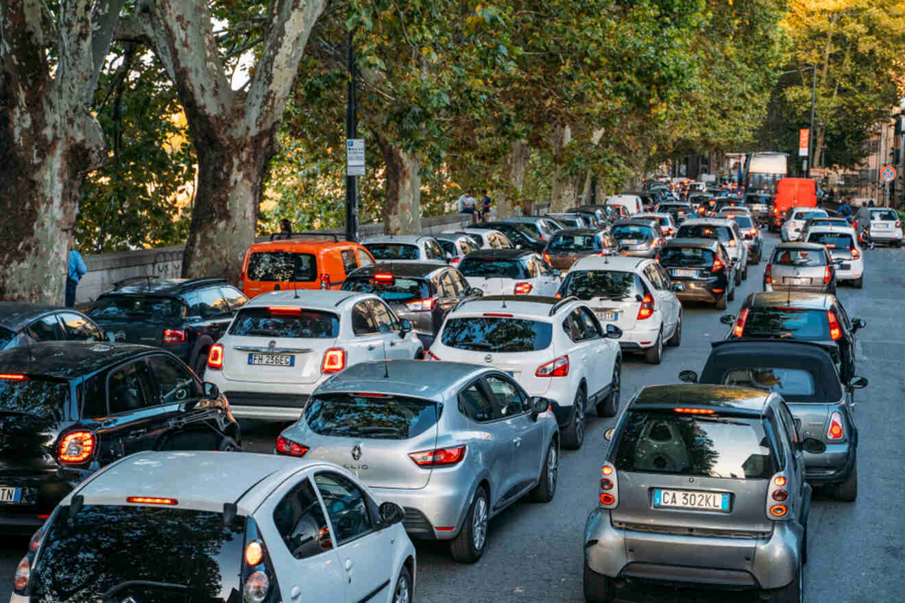 A busy urban street filled with tightly packed traffic, including various makes and models of cars, under large leafy trees.