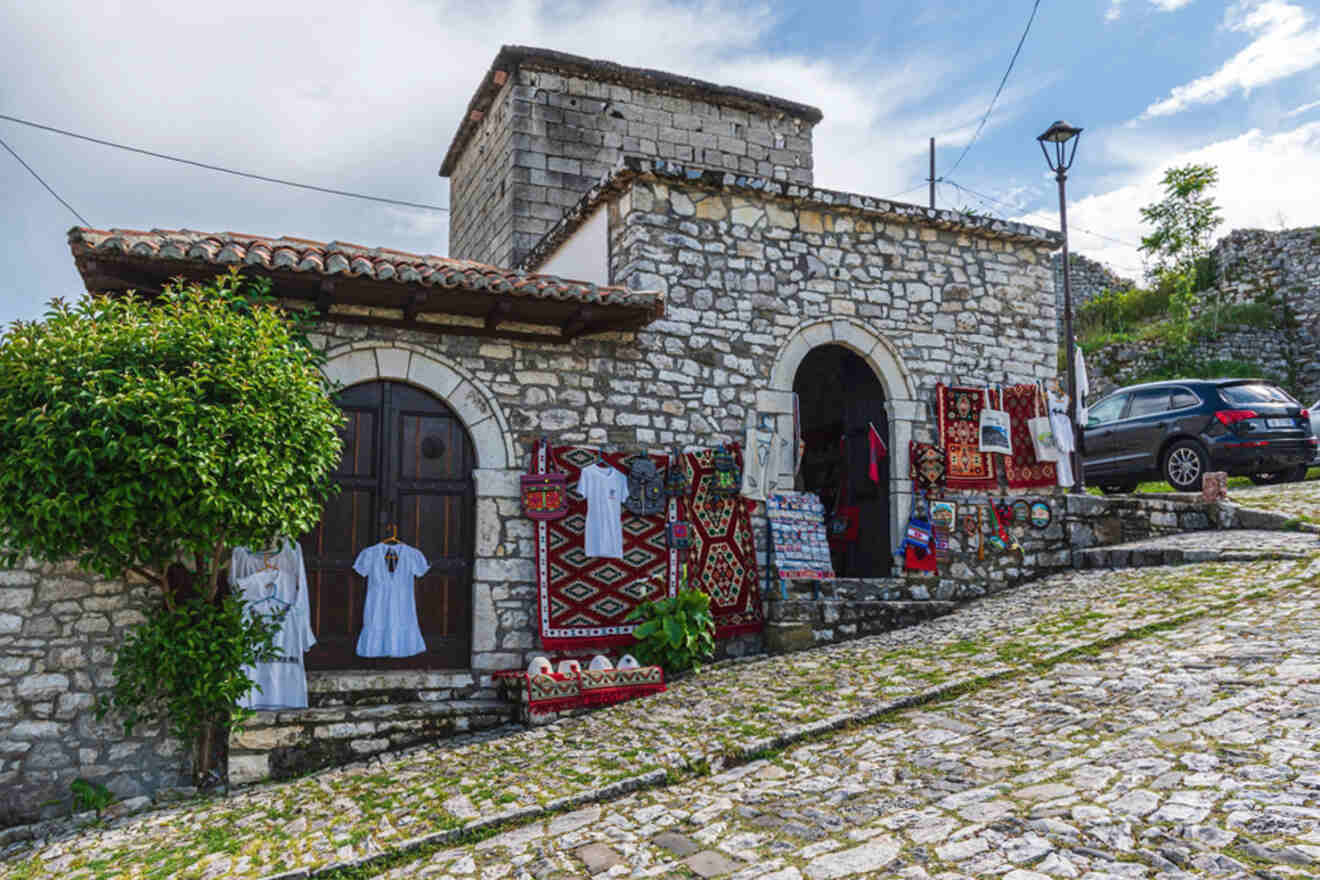 A stone building displays traditional woven rugs and garments outside. An arched wooden door is partly visible. A black car is parked on the cobblestone street nearby.