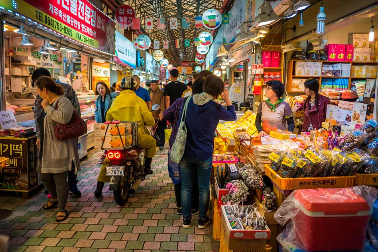 A busy indoor market with people shopping at various stalls selling food and goods. A motorbike is seen navigating through the market. Bright signs and food products are displayed prominently.