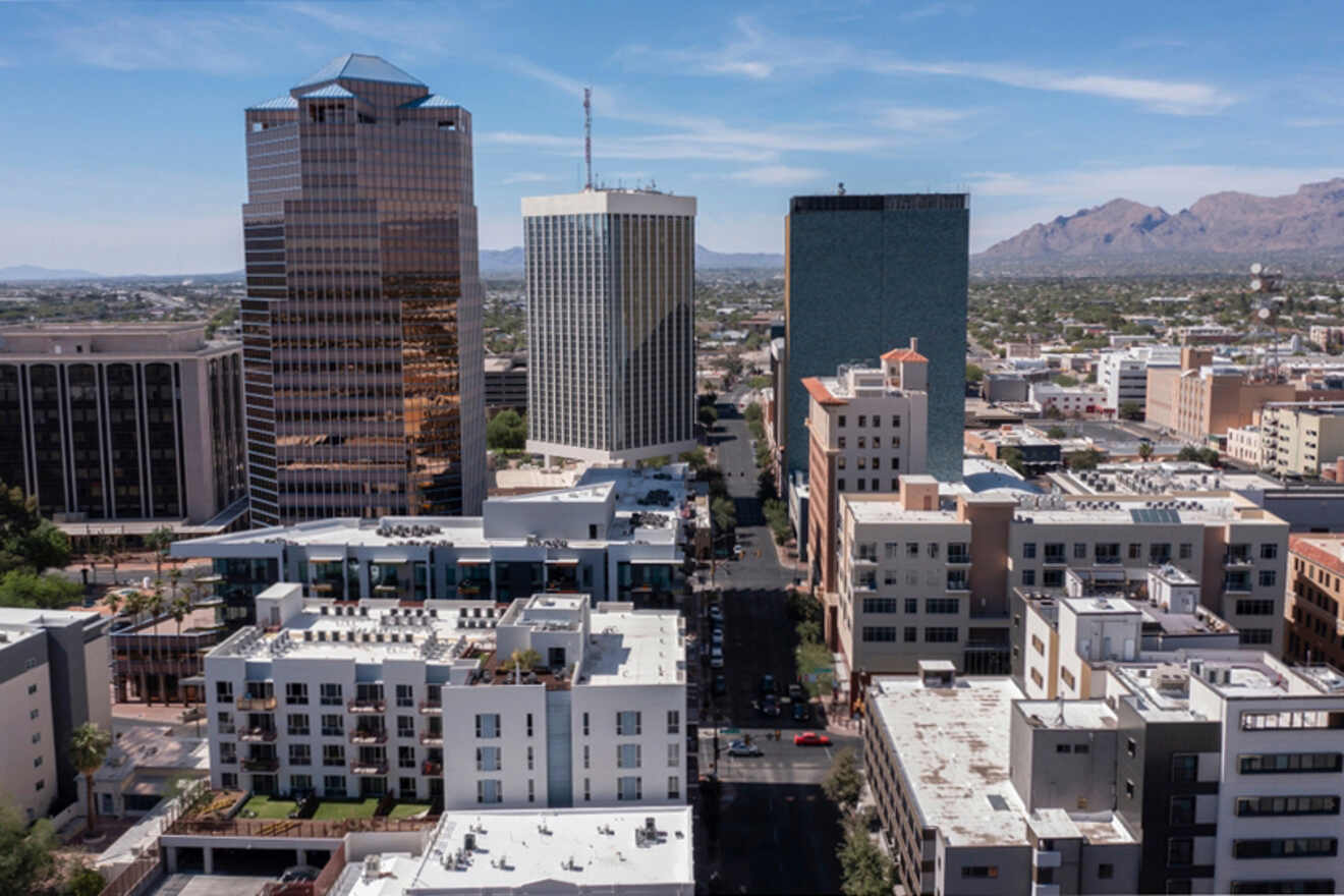 Aerial view of a cityscape featuring several high-rise buildings and surrounding structures with a mountainous backdrop under a clear sky.