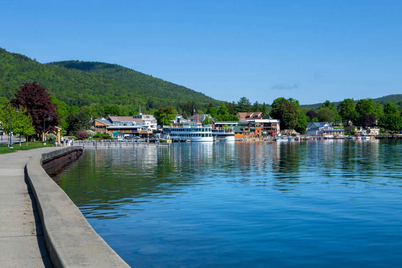 A scenic waterfront view showing a small town with buildings, a boardwalk, and boats docked at a pier, surrounded by green hills under a clear blue sky.