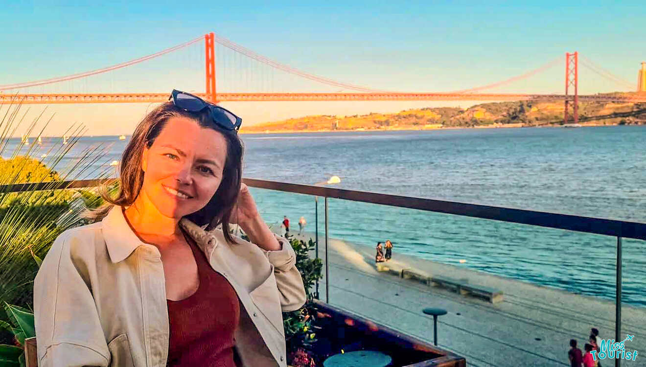 The writer of the post sits outdoors with a view of a large red suspension bridge reminiscent of those in Portugal, overlooking shimmering water under a clear blue sky.