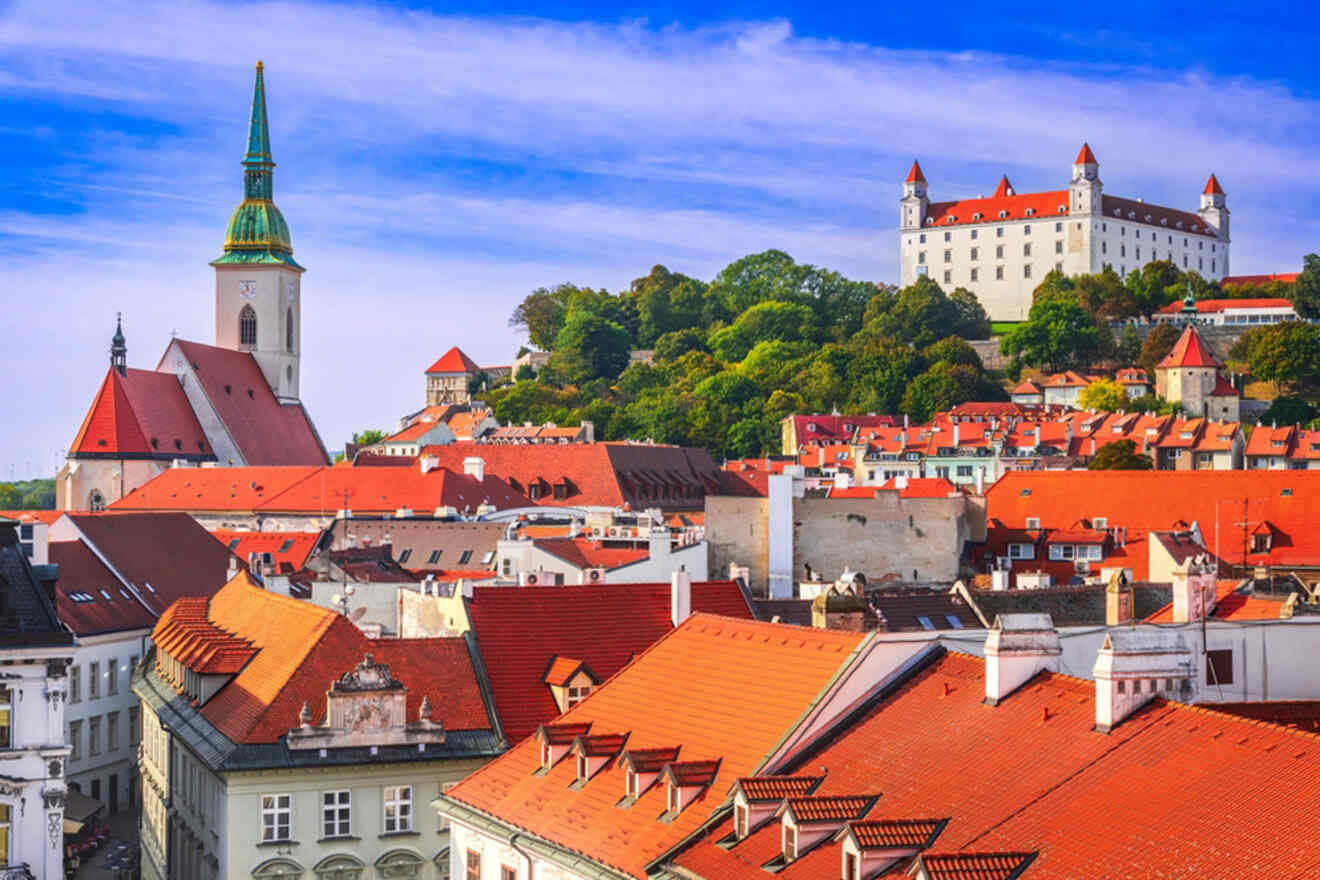 Panoramic view of Bratislava featuring St. Martin's Cathedral, red-roofed buildings, and Bratislava Castle situated atop a hill in the background under a blue sky.