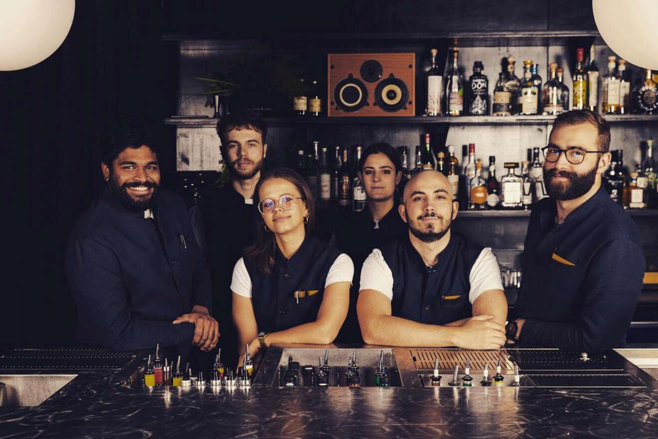 A group of six bartenders stand behind a bar with bottles on the shelves, smiling in a dimly lit setting.