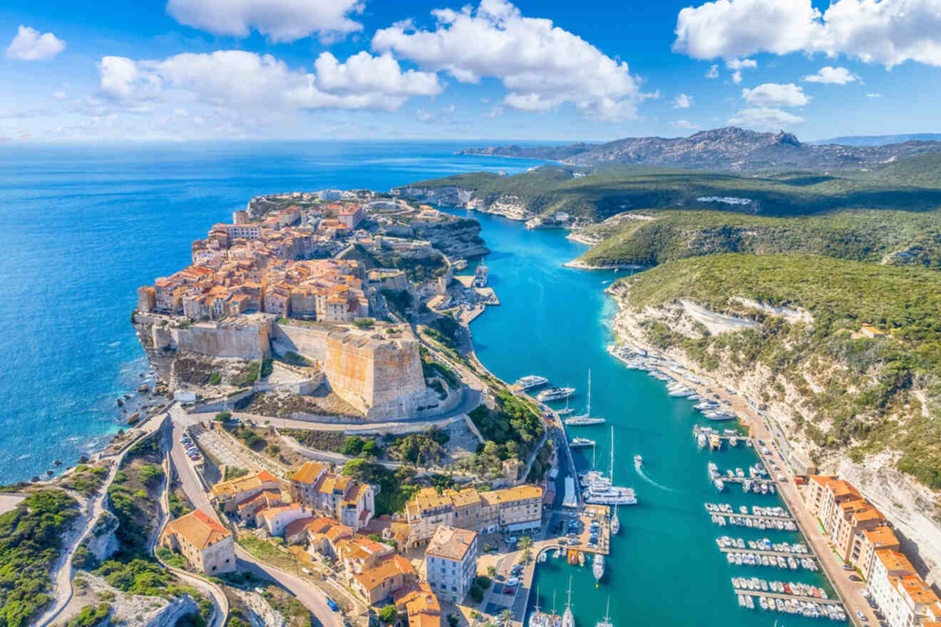 An aerial view of a coastal town with terracotta-roofed buildings perched on cliffs, surrounded by turquoise waters and lush green hills in the distance.