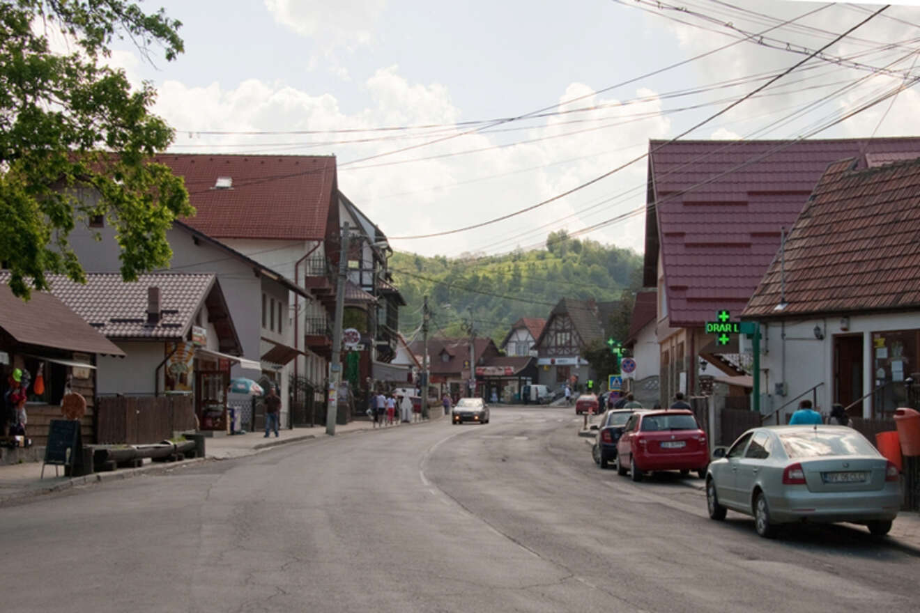 A street in a small town with several houses, parked cars, scattered pedestrians, and a green pharmacy sign. The background shows a tree-covered hill.