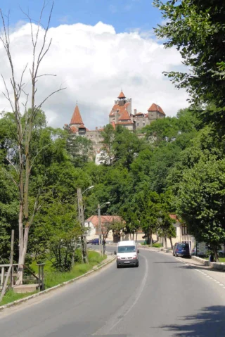 View of a winding road leading to a hilltop castle surrounded by lush greenery and under a partly cloudy sky. A white van is driving on the road.