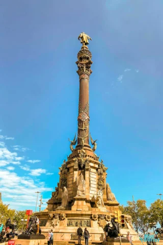 A tall monument with a column and a bronze statue on top, surrounded by decorative sculptures and figures, stands under a clear sky, with people gathered around its base.