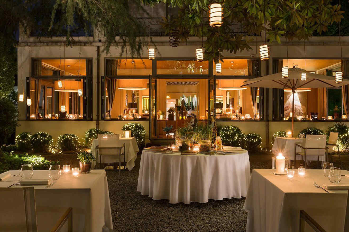 An outdoor dining area at night with elegantly set tables, ambient lanterns, and soft string lights along the building and shrubs.