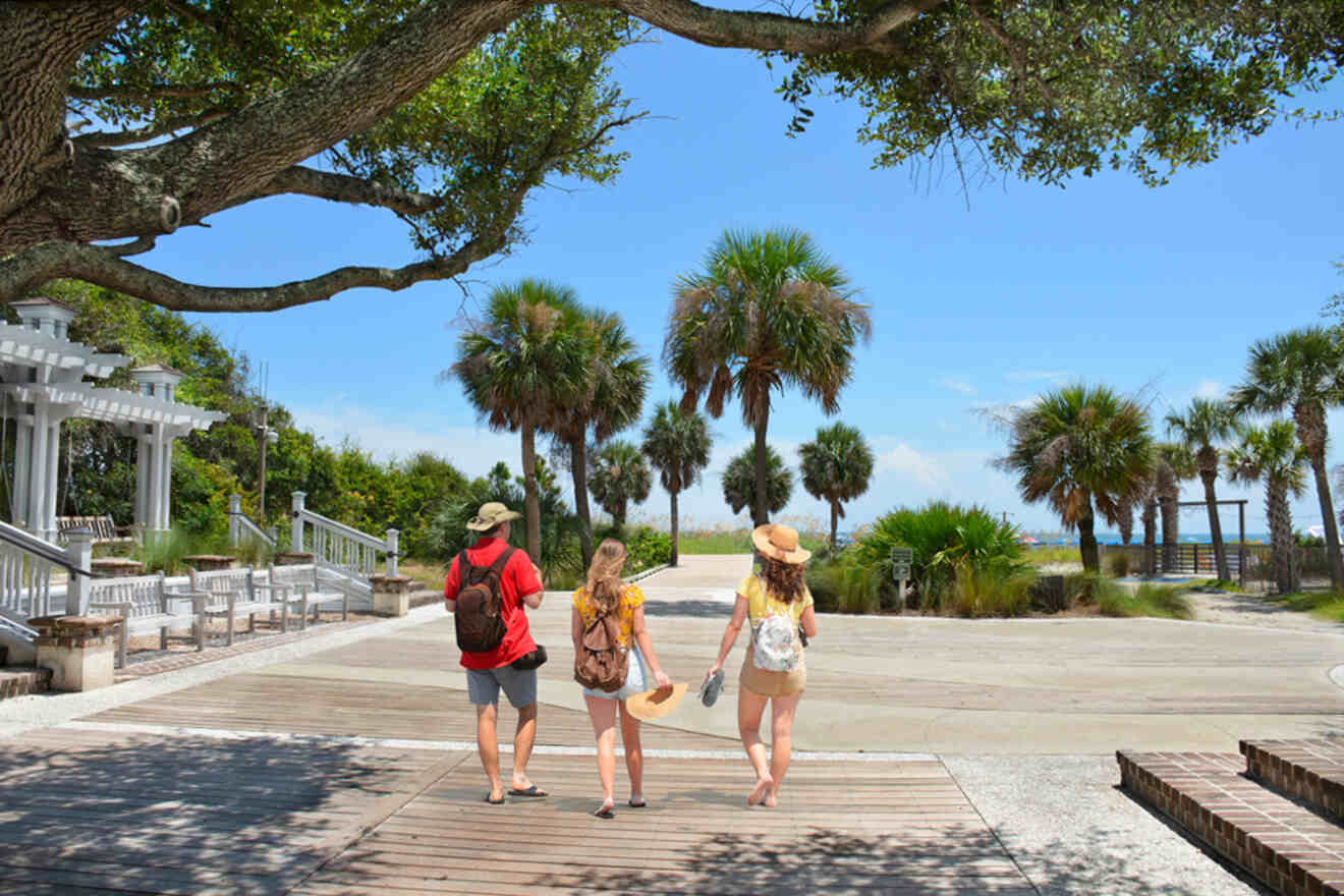 Three people with backpacks and hats walk along a wooden boardwalk towards a beach, surrounded by palm trees on a sunny day.