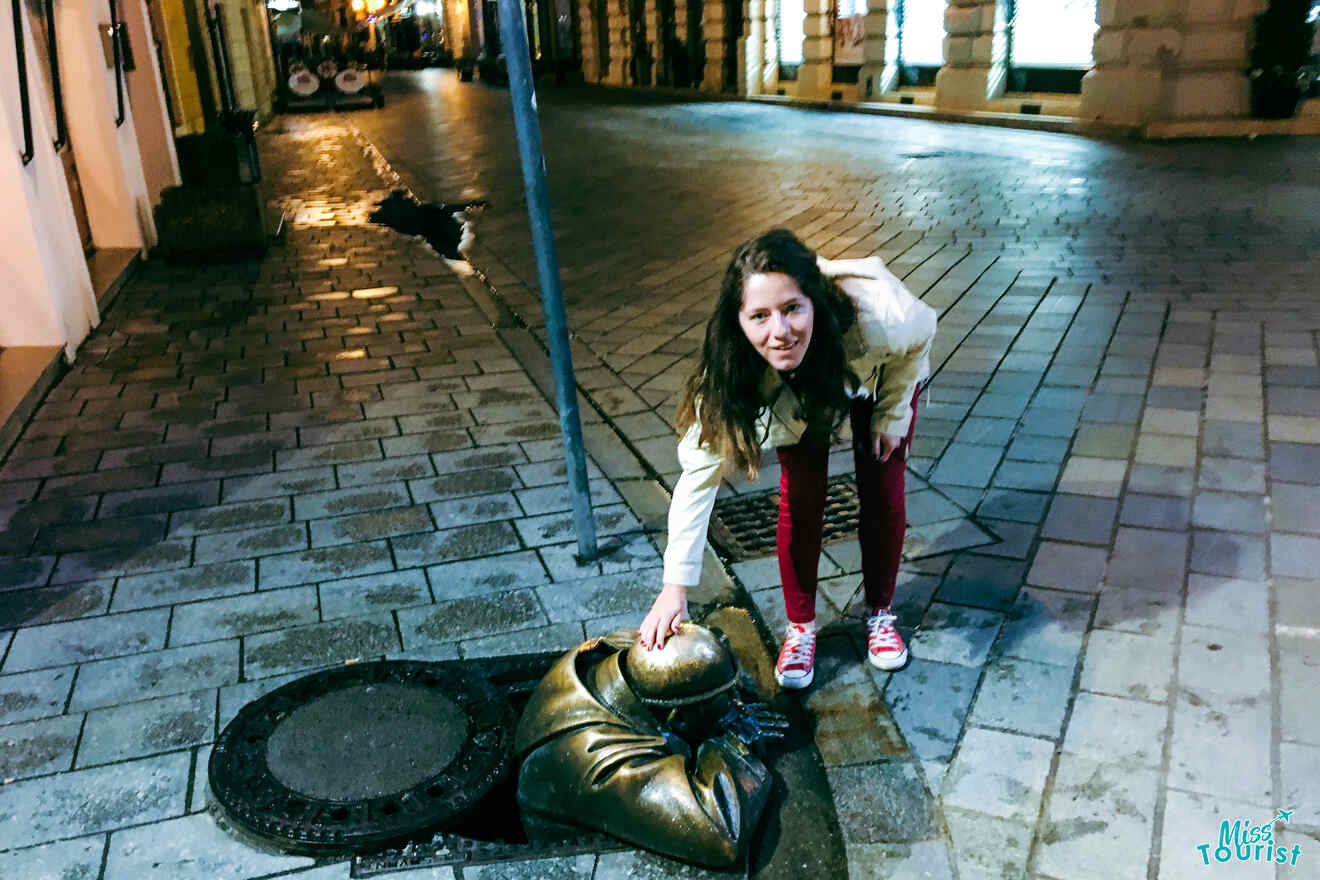 author of the post in a white jacket and red pants touches the head of a bronze statue emerging from a manhole on a cobblestone street at night.