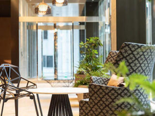 Modern indoor seating area with patterned chairs around a white table. The space features plants, large windows, and contemporary pendant lights.