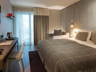 Modern hotel room with a large bed, two bedside lamps, a desk and chair, and a window with sheer curtains leading to a small balcony. The decor is simple with a neutral color palette.