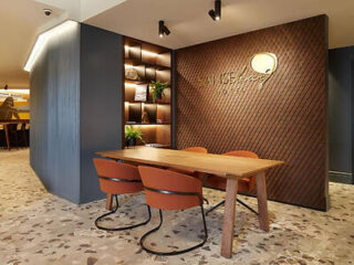 A modern office space featuring a wooden desk with three chairs, a wall shelf with books and plants, and a logo on the back wall. The floor is tiled with a speckled pattern.