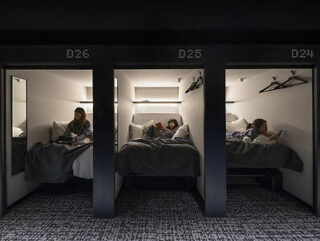 Three individuals are resting in separate, numbered sleeping pods. Each pod has a bed, a small light, and a hanger with clothes. The ambiance is dimly lit and quiet.