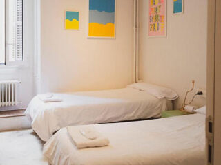 A room with two single beds, each with white bedding and a folded towel. The walls feature colorful framed artworks. There is a small window with white shutters on the left side.