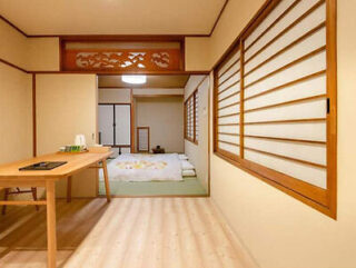 A minimalist Japanese-style room with a wooden floor, sliding shoji doors, a low table with a teapot, and a tatami mat area in the background with futons.