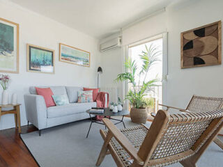 A bright living room with a gray sofa, wooden chairs, potted plants, and framed artworks on the walls. A large window lets in natural light, and a floor lamp stands beside the sofa.