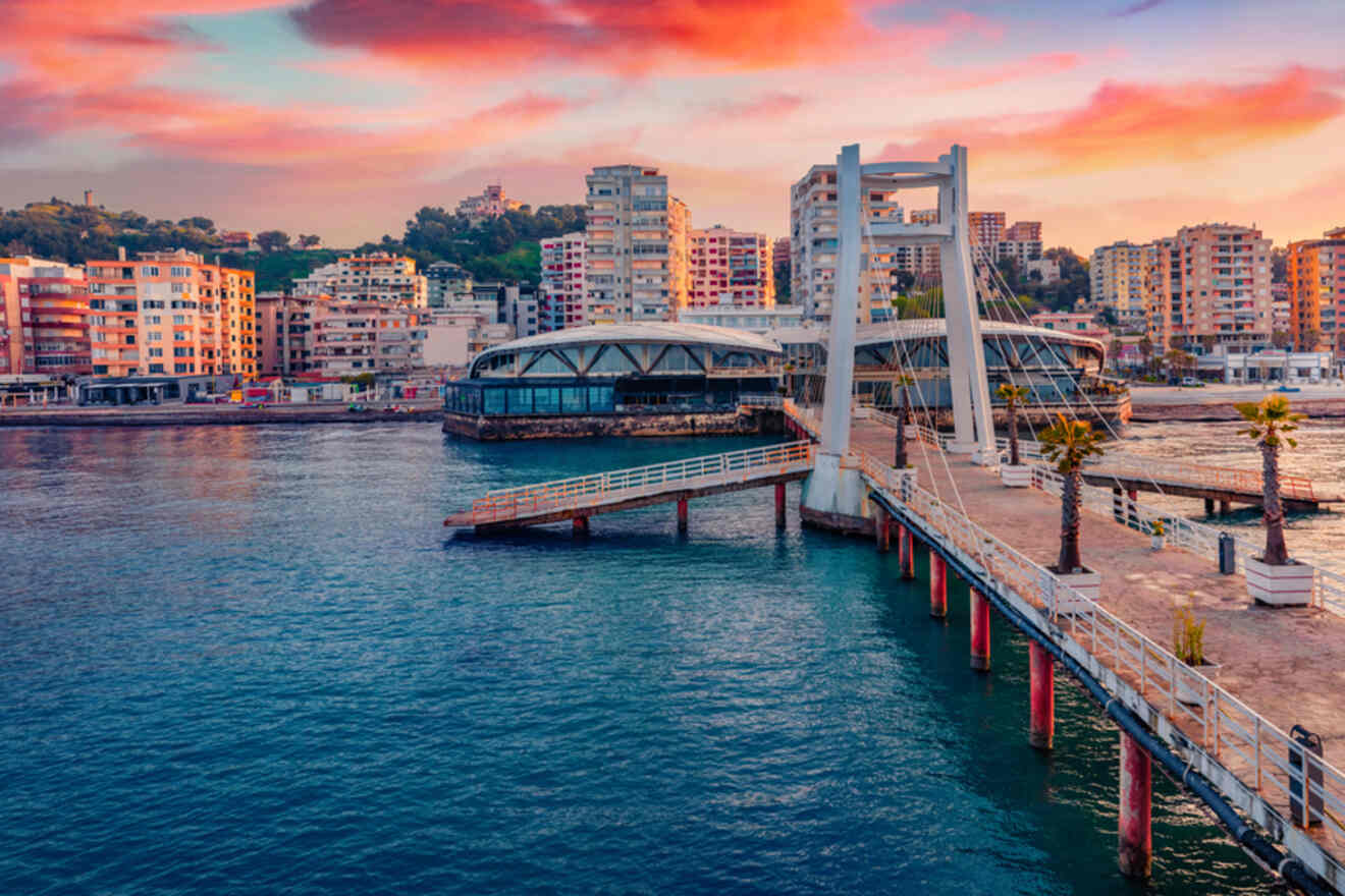 A waterfront scene in Durrës, Albania, with modern buildings and a pedestrian bridge at sunset, casting a soft pink glow over the sky and reflecting on the water.