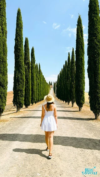 The writer of the post walks down a narrow stone pathway beside an old building, with a view of rolling Tuscan hills and tall cypress trees in the background