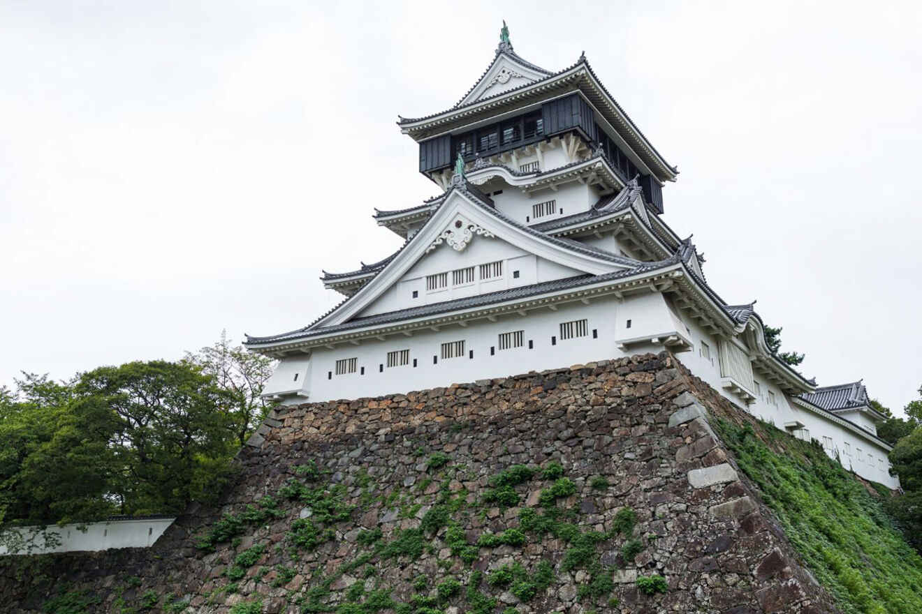 A traditional Japanese castle with white walls, dark-tiled roofs, and a stone base surrounded by greenery under a cloudy sky.