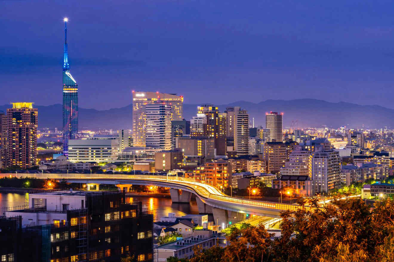 A cityscape at dusk featuring illuminated buildings, a prominent tower, and a brightly lit bridge crossing over water with distant mountains in the background.