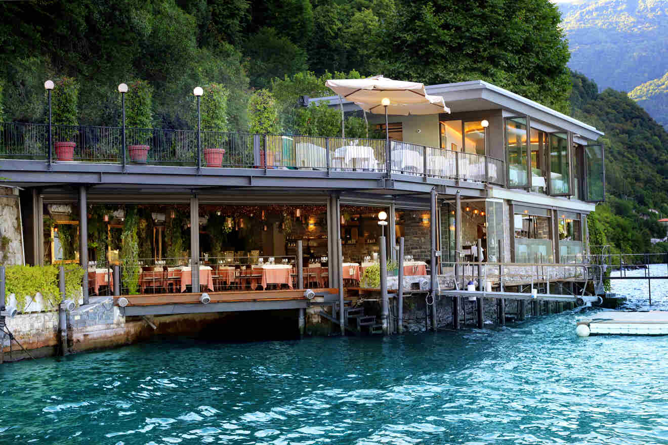 A modern, glass-walled restaurant with outdoor seating overlooks a clear blue lake, surrounded by lush green hills.