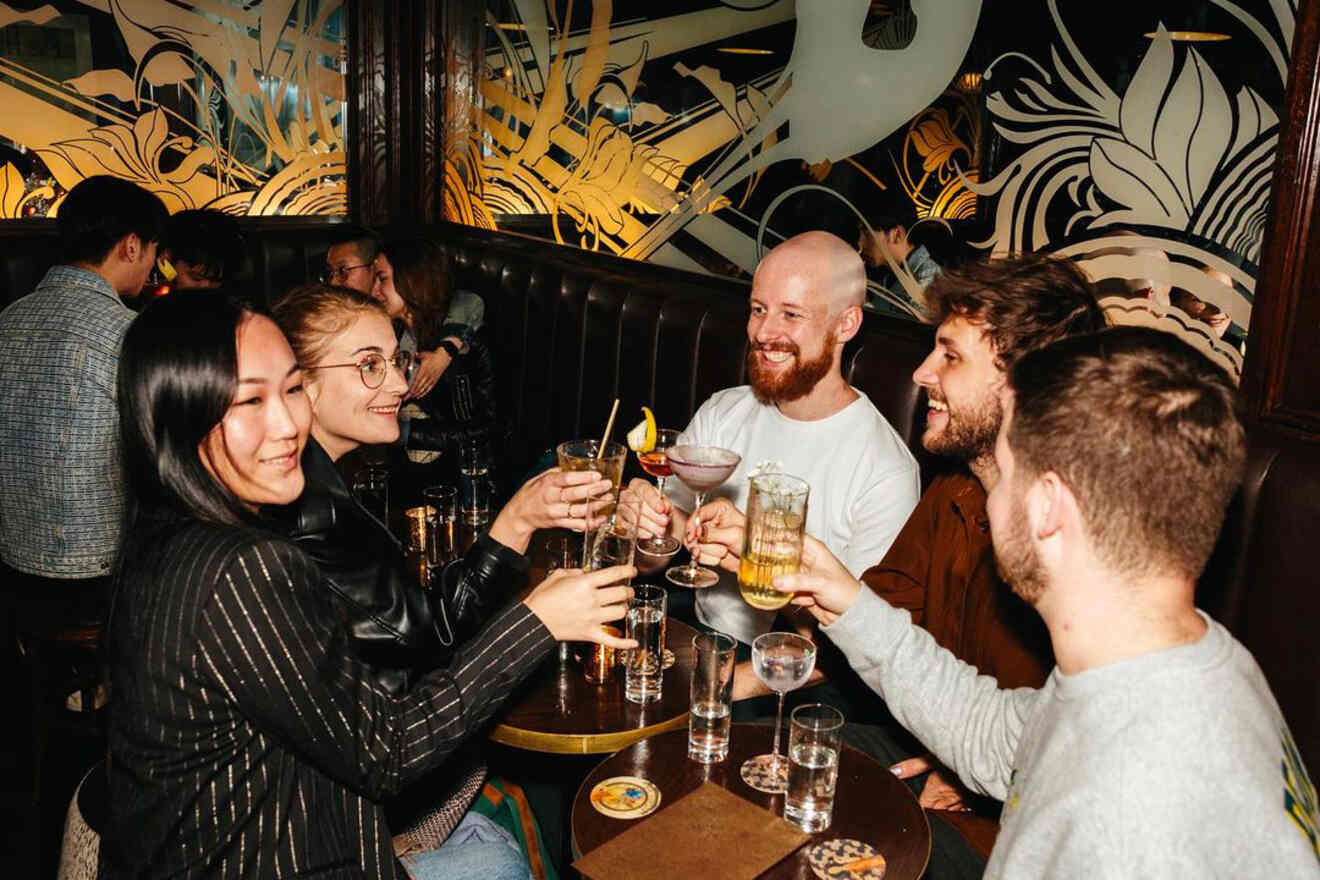 A group of friends seated around a table in a cozy booth clink their drinks together, smiling and enjoying the night out.