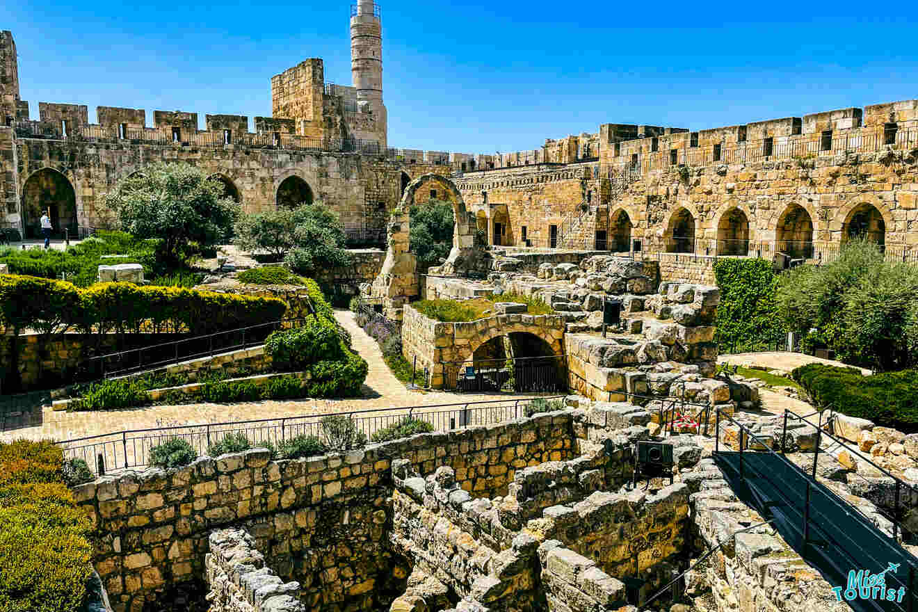 A historic stone fortress with towering walls and arches, surrounded by green foliage. Stone pathways and ruins are visible in the foreground, with a tower rising in the background.