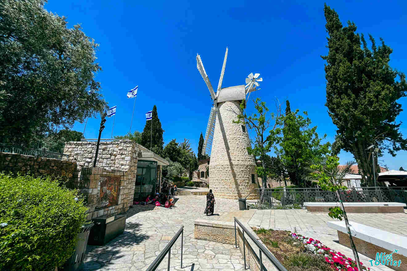 A stone windmill stands in a sunny outdoor setting with flags fluttering nearby, surrounded by trees and visitors. A walkway and a small garden are in the foreground.