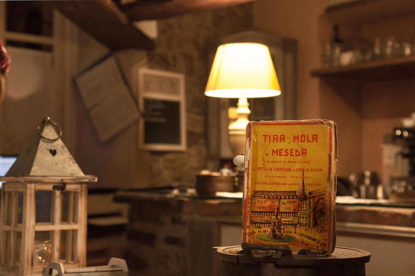 A warmly lit room features a rustic setting with a lit table lamp. In the foreground, an old box with Italian text is displayed, and nearby is a glass lantern. Shelves with dishes are seen in the background.