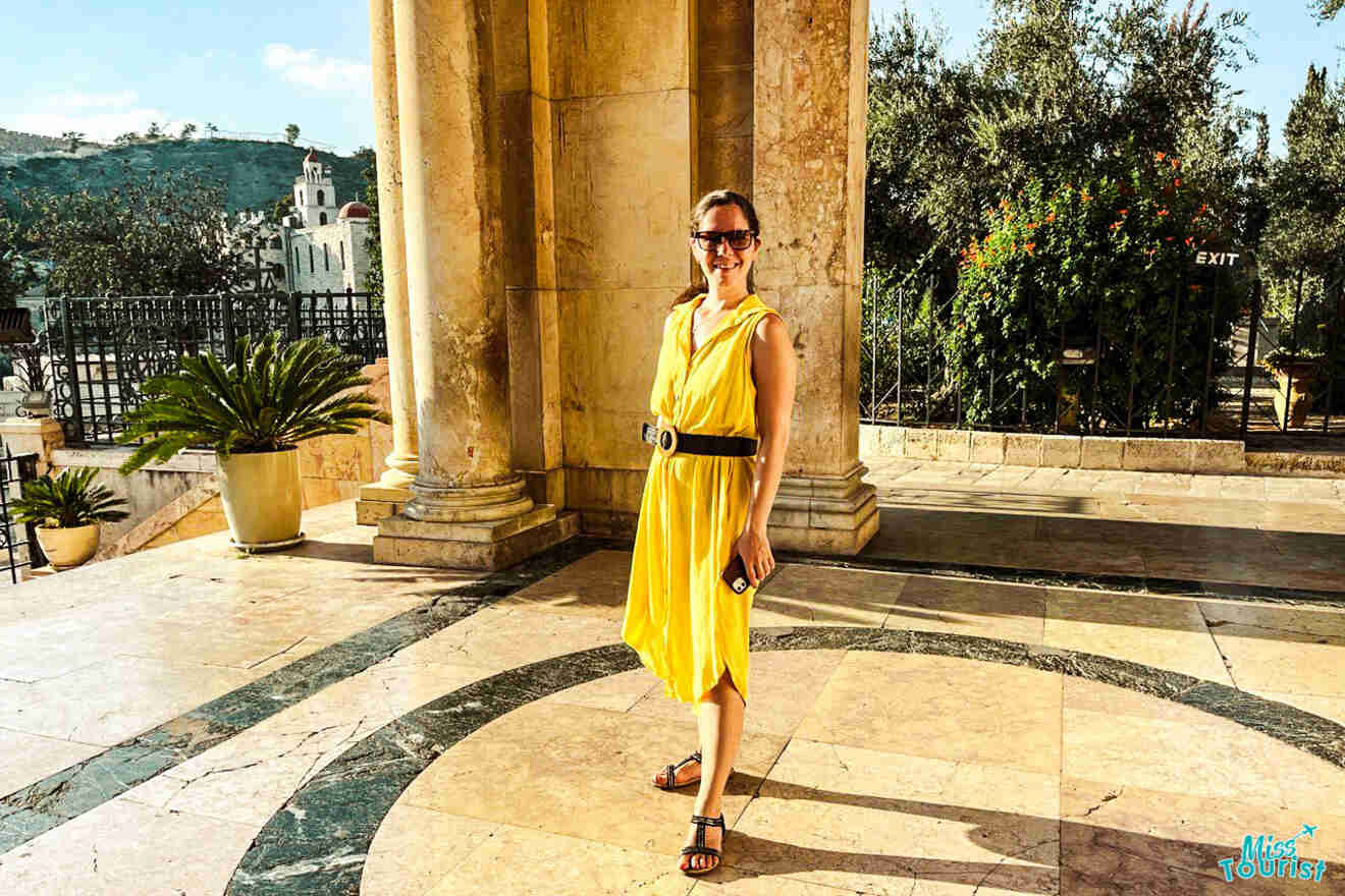 The writer of the post in a yellow dress stands in front of a large stone building with columns. There are potted plants and trees in the background. The person is smiling and holding a phone.