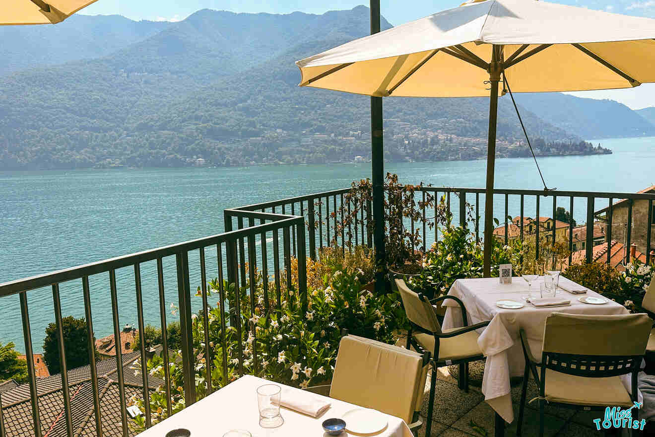 Outdoor restaurant terrace overlooking a serene lake surrounded by mountains, with tables and chairs set under umbrellas, and nearby rooftops visible in the foreground.