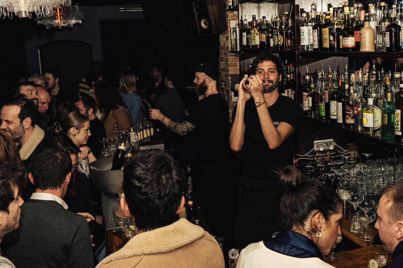 A bartender shakes a cocktail in a crowded, dimly lit bar filled with patrons enjoying drinks and conversation.