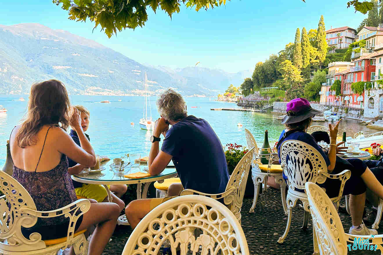 A group of people sit at a terrace overlooking a scenic lake with boats and mountainous landscape in the background. The terrace features white ornate chairs and tables.