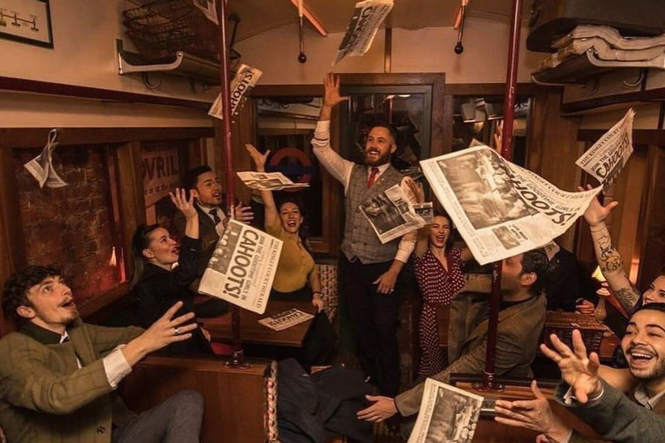 A group of people in vintage clothing celebrate enthusiastically inside a retro-styled bar, tossing up newspapers with "Cahoots!" written on them.