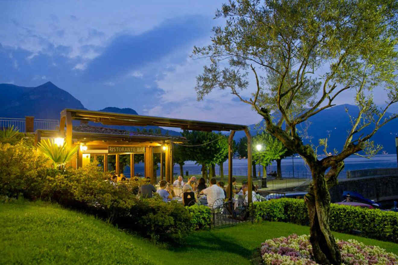 Outdoor restaurant with patrons dining in the evening, surrounded by greenery and a view of mountains and water in the background.