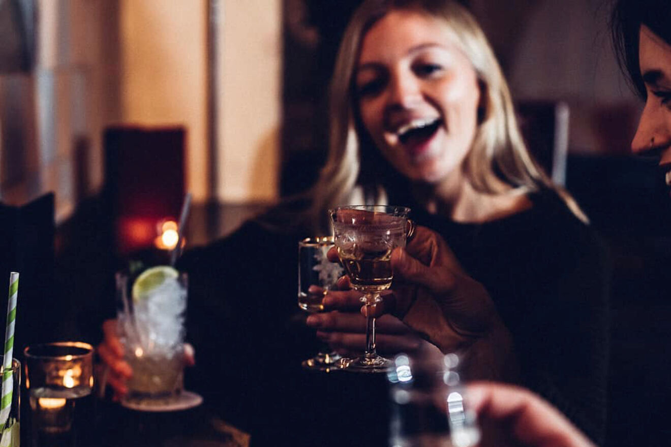 A woman with a big smile raises a glass while toasting with friends in a warmly lit bar, radiating joy and laughter.