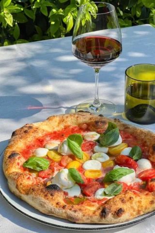 A fresh pizza topped with mozzarella, tomatoes, and basil sits on a table next to a glass of red wine and a green glass in an outdoor setting.