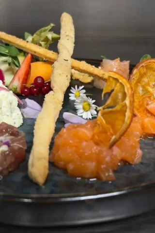 A plate featuring a variety of foods, including breadsticks, diced salmon, dehydrated orange slices, berries, edible flowers, and assorted greens.