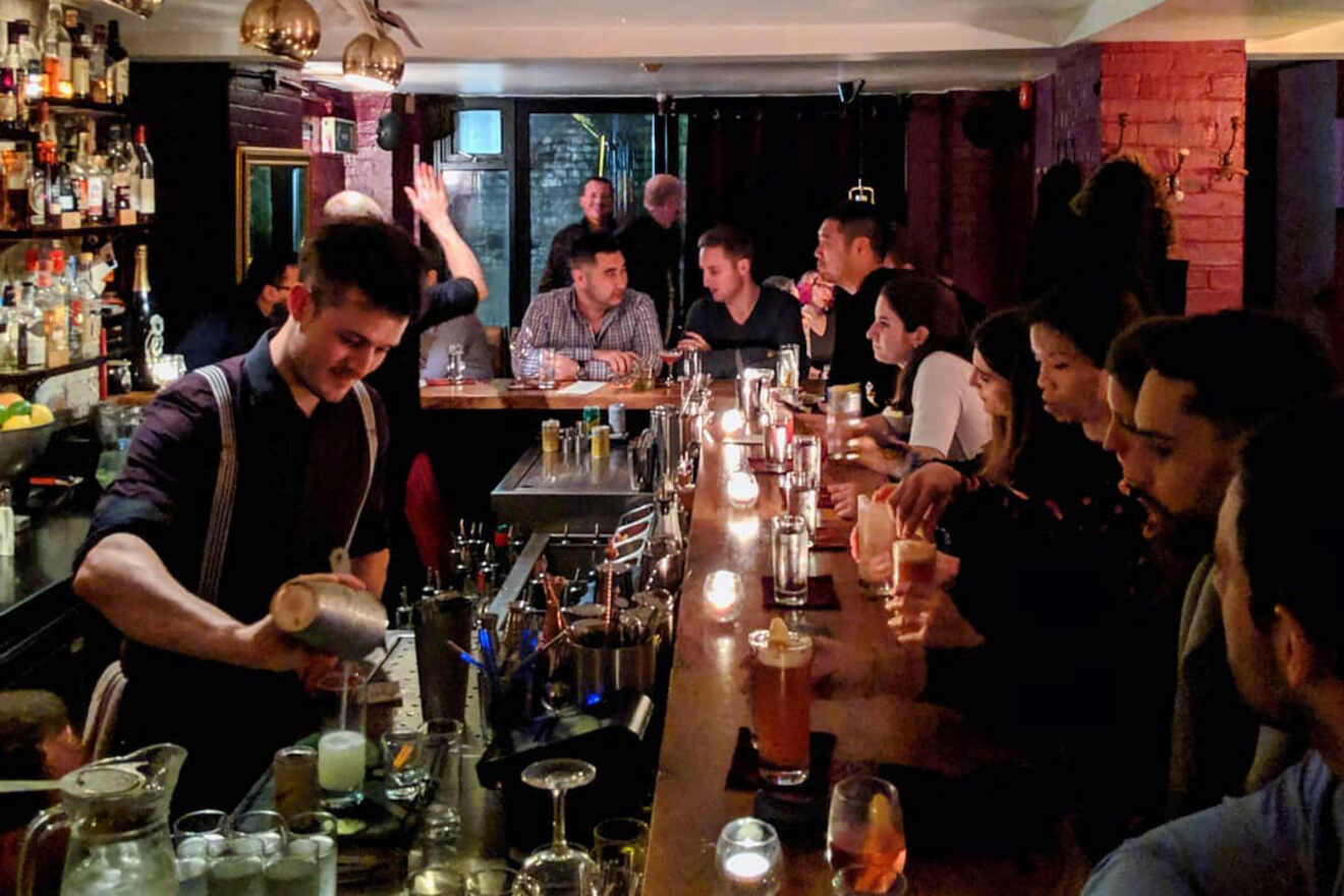 A bartender pours a cocktail for patrons sitting at a long, dimly lit bar, filled with lively conversation and warm ambiance.